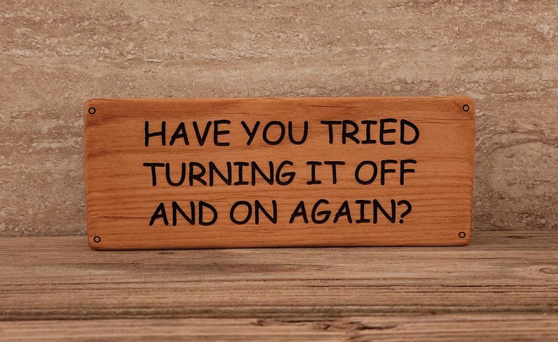 May include: A wooden sign with the text "Have you tried turning it off and on again?" in black lettering.