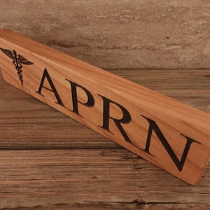 May include: A wooden desk nameplate with the text "APRN" and a medical symbol engraved on the left side.