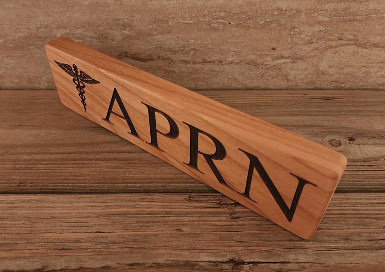 May include: A wooden desk nameplate with the text "APRN" and a medical symbol engraved on the left side.