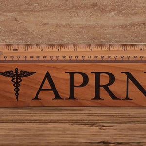 May include: A wooden sign with the text "APRN" and a medical caduceus symbol. The sign is approximately 6 inches long.