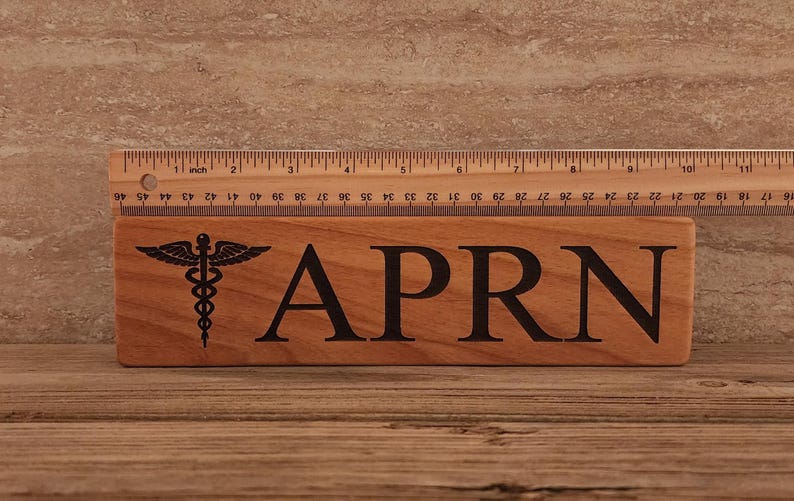 May include: A wooden sign with the text "APRN" and a medical caduceus symbol. The sign is approximately 6 inches long.