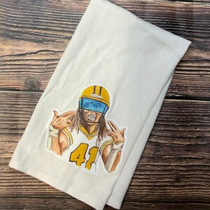 May include: A white kitchen towel with a cartoon illustration of a football player wearing a yellow helmet with the word "Saints" on it. The player is wearing a yellow jersey with the number "41" on it. The player is making a hand gesture.