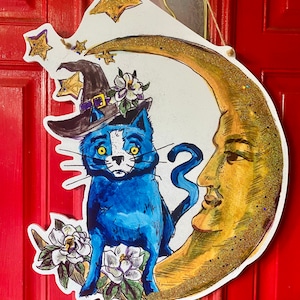 May include: A blue cat wearing a witch's hat sits on a crescent moon with a human face. The moon is decorated with gold glitter. There are stars in the background.