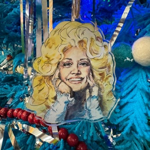 May include: A clear plastic Christmas ornament featuring a watercolor illustration of a woman with blonde hair and a white shirt. The ornament is hanging on a blue Christmas tree.