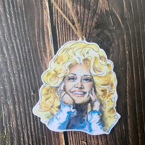 May include: A hand-painted wooden ornament featuring a portrait of a woman with blonde hair and a blue shirt. The ornament has a string for hanging.