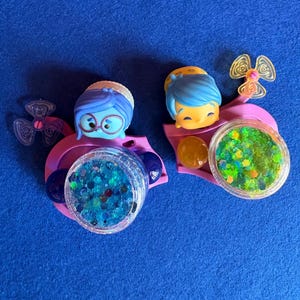 May include: Two character-themed accessories with glitter-filled containers. One features a blue-haired character with glasses, the other a yellow-skinned character with blue hair. Both are set on a pink base with a propeller design.