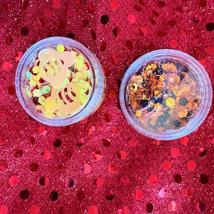 May include: Two clear plastic containers filled with glitter. One contains yellow and gold glitter with a bee-shaped cutout. The other contains orange, black, and gold glitter. The containers sit on a red sequined surface.