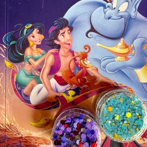 May include: An illustration from the Disney movie Aladdin, featuring Aladdin, Jasmine, the Genie, and Abu. Two clear jars of glitter are in the foreground, one with purple and red glitter, the other with blue and yellow glitter.