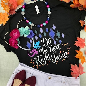 May include: Black t-shirt with a white graphic that says "Do the Next Right Thing" with a blue and purple lizard design. The shirt is laying on a white surface with a pair of red shoes and a floral headband.