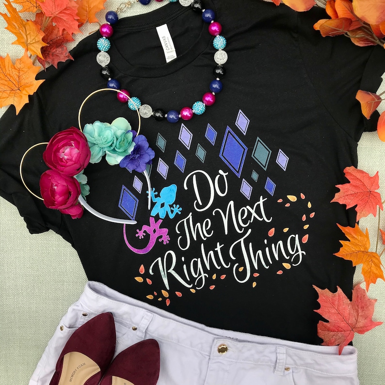 May include: Black t-shirt with a white graphic that says "Do the Next Right Thing" with a blue and purple lizard design. The shirt is laying on a white surface with a pair of red shoes and a floral headband.