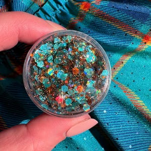 May include: A small, clear container filled with iridescent glitter in shades of teal, copper, and orange. The glitter is in various geometric shapes, reflecting light. The container is held against a teal and plaid fabric background.