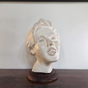 Peut inclure: Buste clair d'une femme aux cheveux courts et coiffés, monté sur une base en bois brun foncé. La sculpture a une expression sereine, avec les lèvres légèrement entrouvertes. Le buste est placé sur un fond neutre.