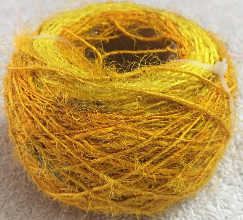 May include: A skein of yellow and gold silk yarn, wound into a ball shape. The yarn is soft and fluffy, with a slightly textured surface.