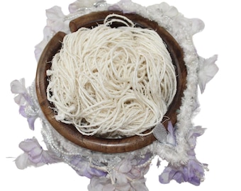 May include: A wooden yarn bowl holds a large ball of off-white yarn. The bowl is surrounded by a decorative arrangement of white and lavender fabric, flowers, and silver accents. The yarn is tightly wound.