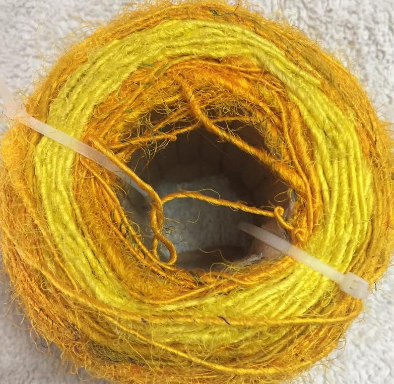 May include: A skein of yellow and orange yarn, tied with a white plastic tie. The yarn is made of natural fibers and has a fuzzy texture.