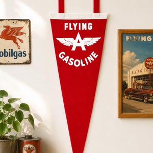 May include: A red triangular felt pennant featuring the white text FLYING GASOLINE and a central white winged letter A logo. The pennant has a white top border and two red fabric tabs at the top for hanging, displaying a vintage-inspired design.