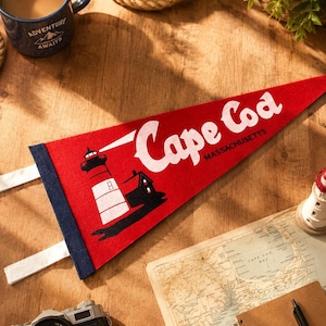 May include: A red felt pennant featuring the white text Cape Cod Massachusetts and a graphic of a lighthouse. The pennant has a dark blue vertical strip on the left side with two white fabric ties attached to the edge for hanging purposes.