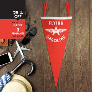 May include: Red pennant with white text that reads "FLYING A GASOLINE" and a stylized wing design. The pennant has a white border and red hanging straps. Other items include sunglasses, a model airplane, and a straw hat.