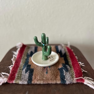 May include: A green ceramic cactus ring holder with a small turquoise ring on it, set on a small white dish. The dish sits on a colorful woven rug with red, brown, and blue patterns. The rug has white fringe.