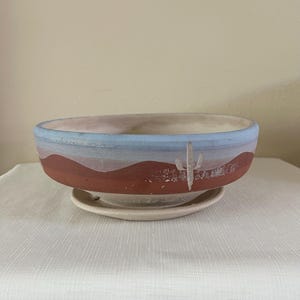 May include: A ceramic planter with a desert landscape design. The planter features a blue rim, a red and white painted landscape, and a cactus illustration. It sits on a matching saucer, perfect for succulents or small plants.