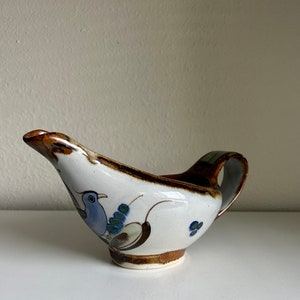 May include: A white ceramic gravy boat with a brown rim and handle. The boat is decorated with a blue bird and green and brown floral designs.