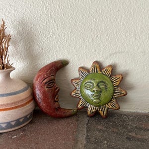 May include: Hand-painted ceramic sun and moon figurines. The sun has a green face with gold rays, and the crescent moon is red. A small vase with a dried plant is on the left.