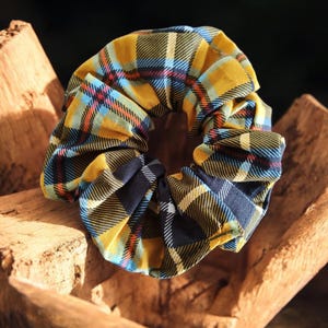 May include: A yellow, blue and black plaid scrunchie hair tie. The scrunchie is made of fabric and is gathered into a circle.