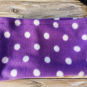 May include: A purple fleece neck warmer featuring large white polka dots. The fabric is soft and plush, ideal for cold weather. The neck warmer is folded. This item is a cosy accessory, perfect for keeping warm.