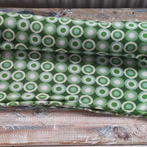 Puede incluir: Una manta polar verde enrollada con un patrón repetido de círculos blancos, verde claro y verde oscuro. La manta es suave y parece estar hecha de un material afelpado. El diseño es moderno y geométrico.