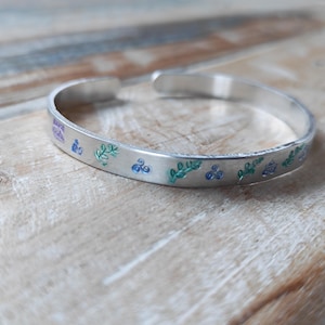 May include: A silver cuff bracelet with a floral and text design. The bracelet features blue and green painted floral accents. The open cuff design allows for easy wear. The bracelet is displayed on a wooden surface.