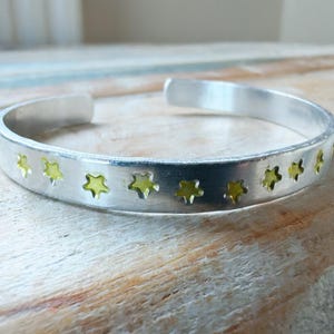 May include: A silver cuff bracelet with star cutouts filled with a yellow-green material. The bracelet is open-ended and rests on a light-colored wooden surface. The bracelet's design is simple and elegant.