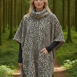 Pode incluir: Uma mulher usando um poncho com estampa de leopardo bege e preto, gola alta e dois bolsos frontais. O poncho é usado sobre uma camisa preta de manga comprida e leggings pretas. O fundo é uma floresta desfocada.