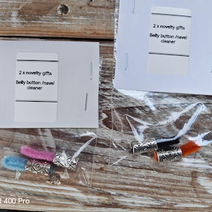 May include: Two novelty belly button/navel cleaners are shown in clear plastic bags. Each cleaner features a decorative silver-coloured bead and a colourful, fluffy cleaning tip. The packaging reads "2 x novelty gifts Belly button/navel cleaner."