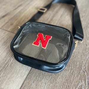 Op de afbeelding: Transparante crossbody tas met een verstelbare zwarte schouderband en een rode viltletter "N" aan de voorkant.