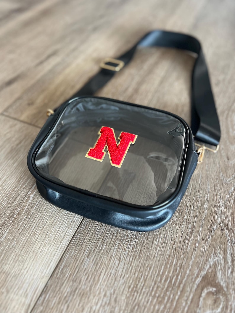 Op de afbeelding: Transparante crossbody tas met een verstelbare zwarte schouderband en een rode viltletter "N" aan de voorkant.