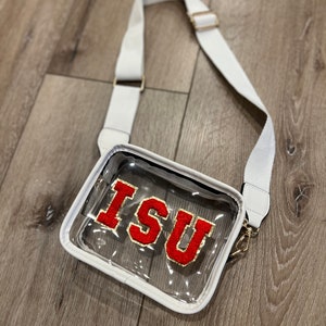 Op de afbeelding: Transparante crossbody tas met een witte band en een rood vilt patch met de letters "ISU" in gouden letters.