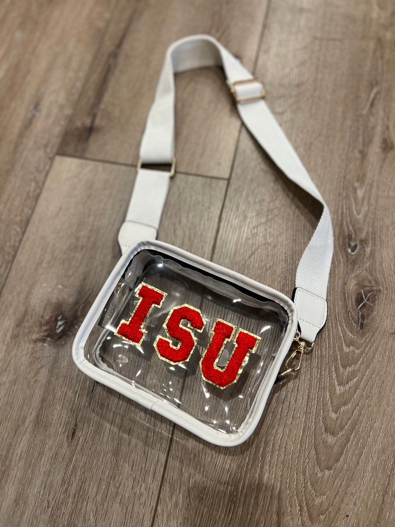 Op de afbeelding: Transparante crossbody tas met een witte band en een rood vilt patch met de letters "ISU" in gouden letters.