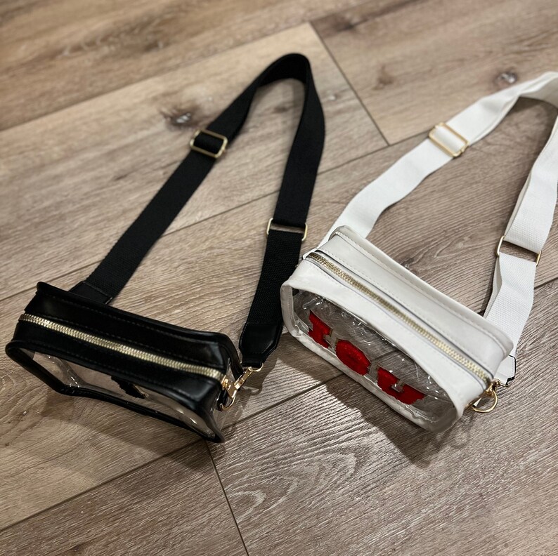 Op de afbeelding: Twee transparante crossbody-tassen met gouden ritsen. E&eacute;n tas heeft een zwarte leren rand en de andere een witte leren rand. De witte tas heeft het woord "YOU" in rode letters op de voorkant.