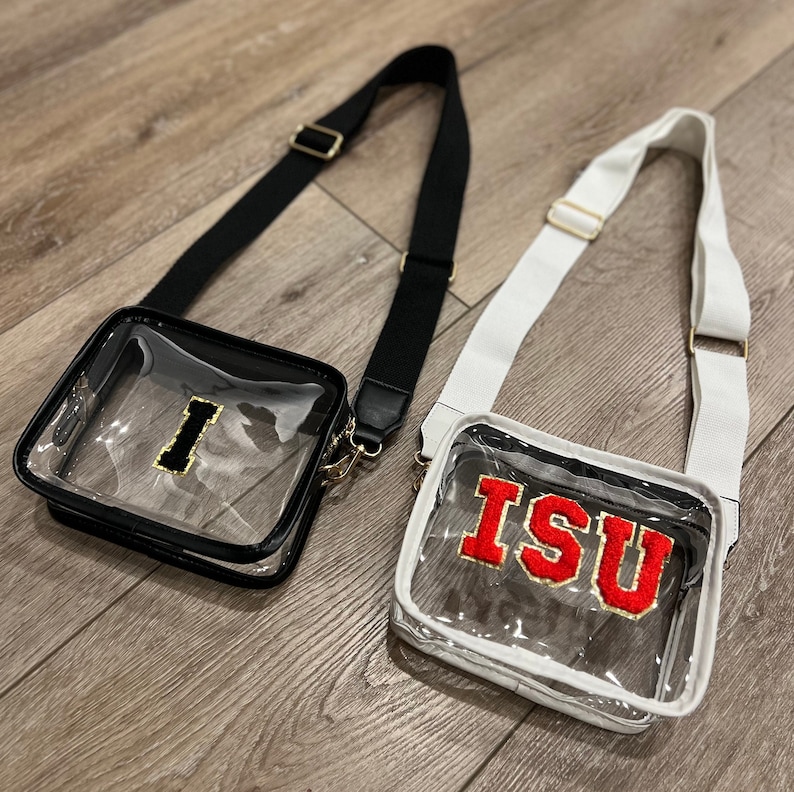 Op de afbeelding: Twee transparante crossbody-tassen met zwarte en witte banden. De zwarte tas heeft een gouden "I" aan de voorkant. De witte tas heeft een rode "ISU" aan de voorkant.