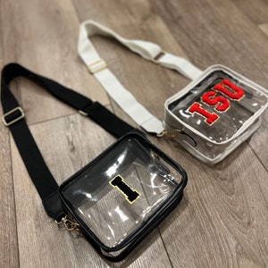 Op de afbeelding: Twee transparante crossbody-tassen met zwarte en witte banden. De tas links heeft een gouden "I" aan de voorkant. De tas rechts heeft een rood "ISU" aan de voorkant.