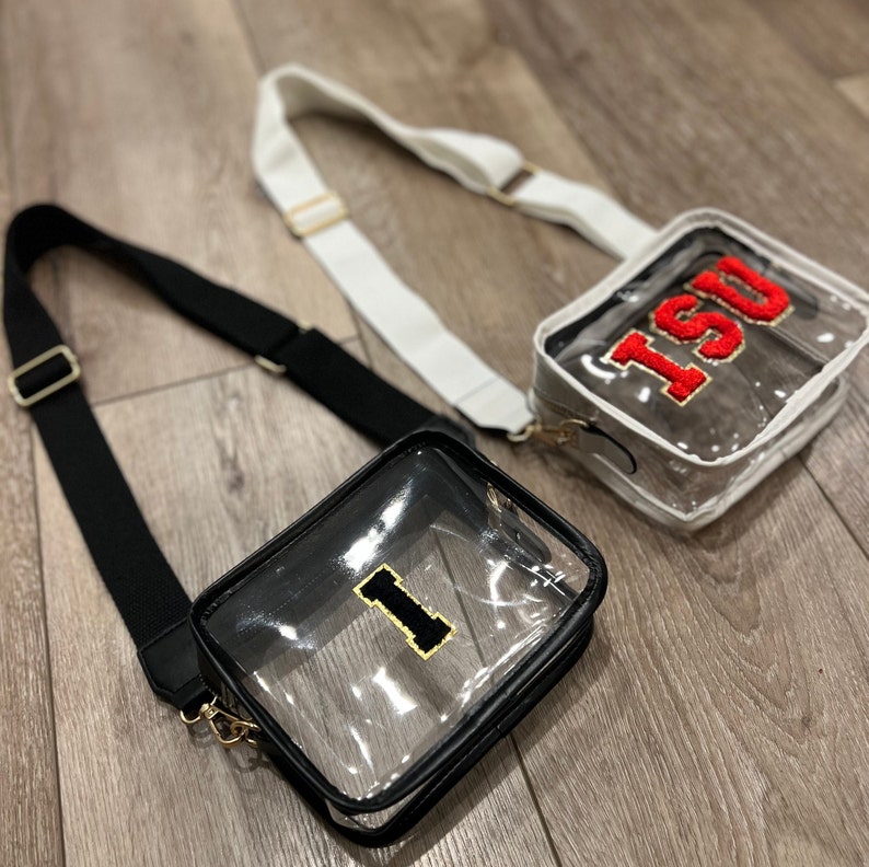 Op de afbeelding: Twee transparante crossbody-tassen met zwarte en witte banden. De tas links heeft een gouden "I" aan de voorkant. De tas rechts heeft een rood "ISU" aan de voorkant.