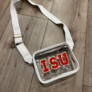 Peut inclure: Un sac à bandoulière transparent en plastique avec une bandoulière blanche et un logo "ISU" rouge.