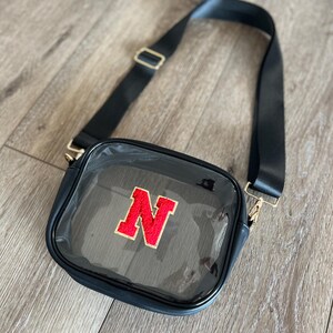 Op de afbeelding: Een transparante crossbody tas van plastic met een zwarte band en een rode viltletter "N" aan de voorkant.