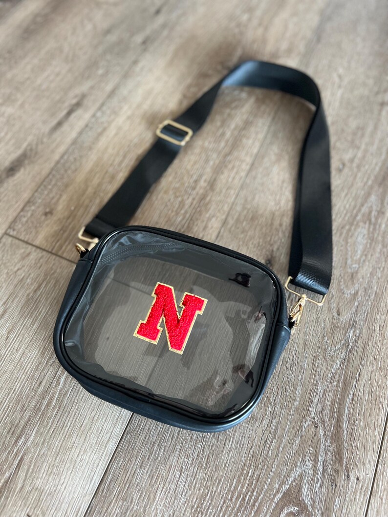 Op de afbeelding: Een transparante crossbody tas van plastic met een zwarte band en een rode viltletter "N" aan de voorkant.