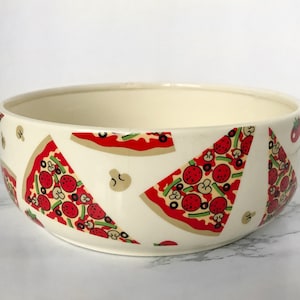 May include: A white ceramic bowl with a pizza slice pattern. The bowl has a red, green, and brown pizza slice design with pepperoni, mushrooms, and green peppers.