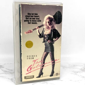 May include: A VHS cassette tape cover with a woman holding a bass guitar. The cover is pink and black with the text "Scenes from The Goldmine" and "Charter".