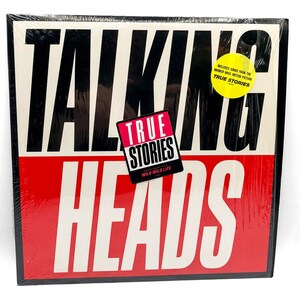 Puede incluir: Una portada de disco de vinilo en blanco y negro con las palabras "Talking Heads" en letras blancas grandes y en negrita. El t&iacute;tulo del &aacute;lbum "True Stories" est&aacute; en una fuente m&aacute;s peque&ntilde;a, rosa con un borde negro. La portada es roja y blanca con una pegatina amarilla que dice "Includes songs from the Warner Bros. motion picture True Stories."