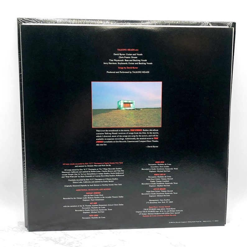 Puede incluir: La contraportada de un disco de vinilo de Talking Heads. El disco se titula "True Stories" y presenta una foto de un peque&ntilde;o edificio verde en un campo. El texto en la contraportada incluye los nombres de los miembros de la banda, el t&iacute;tulo de la canci&oacute;n y los cr&eacute;ditos del &aacute;lbum.