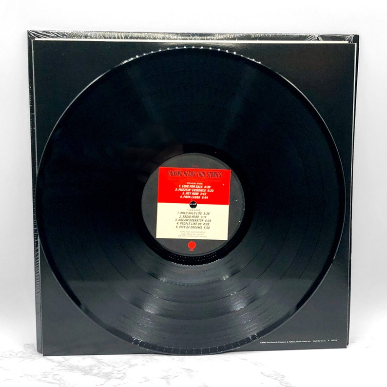 Puede incluir: Un disco de vinilo negro con una etiqueta blanca que dice "Talking Heads True Stories". La etiqueta enumera las canciones del disco, incluyendo "Wild Wild Life", "Radio Head", "People Like Us" y "City of Dreams".