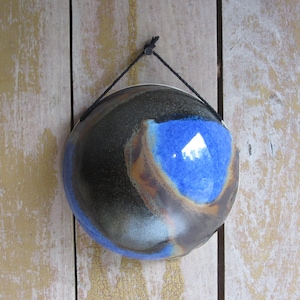 May include: A circular ceramic wall hanging featuring a glossy glaze with a deep black base, a vibrant cobalt blue center, and subtle brown accents. The piece is suspended by a thin black cord attached to the top, creating a smooth, reflective surface.